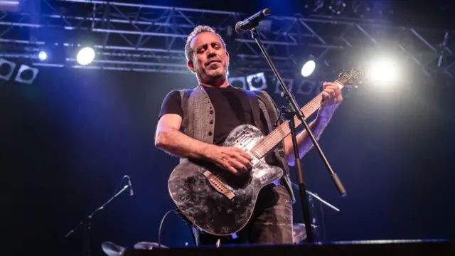 Haluk Levent sahnede şarkı söyleyip gitar çalıyor. Üzerinde siyah bir tshirt ve yelek, elinde siyah bir elektrogitar var.
